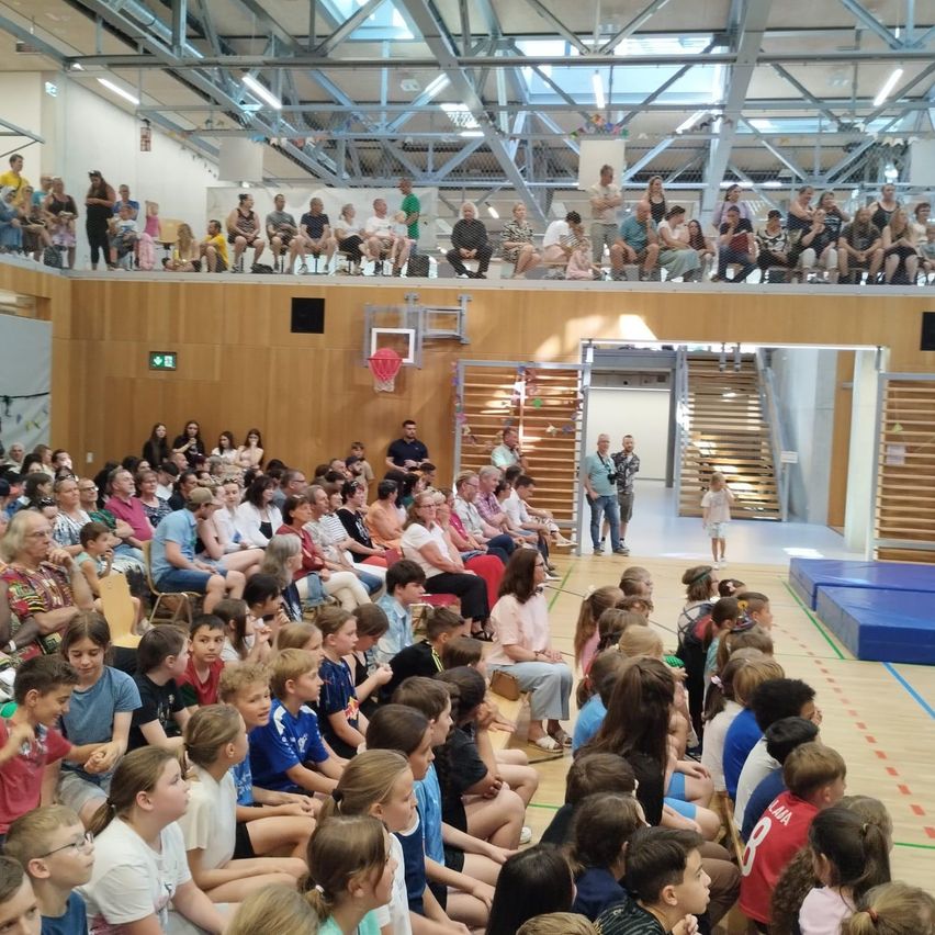 Eine große Menschenmenge sitzt auf Bänken in einer Turnhalle. Einige sitzen auf dem Boden, andere auf der Empore. Der Raum hat einen Basketballkorb, eine blaue Matte und eine Treppe. Eine Person steht in der Nähe des Eingangs.