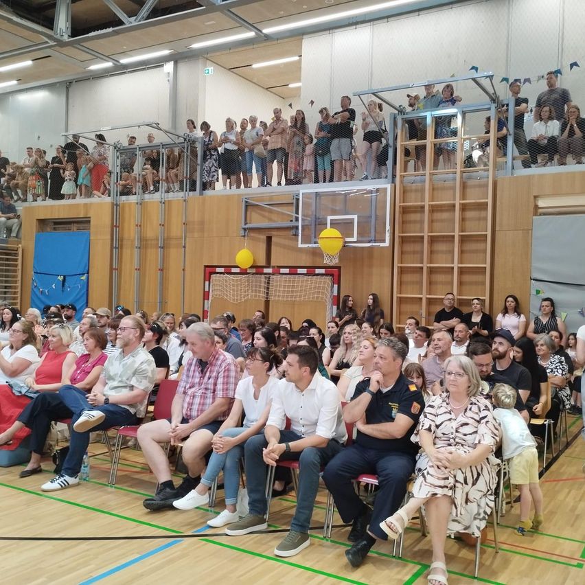 Eine große Menschenmenge versammelt sich in einer Turnhalle und schaut sich ein Sportereignis an. Zuschauer sitzen auf Stühlen und Tribünen, während andere auf der Galerie stehen. Ein Basketballkorb und ein gelber Ball sind sichtbar.