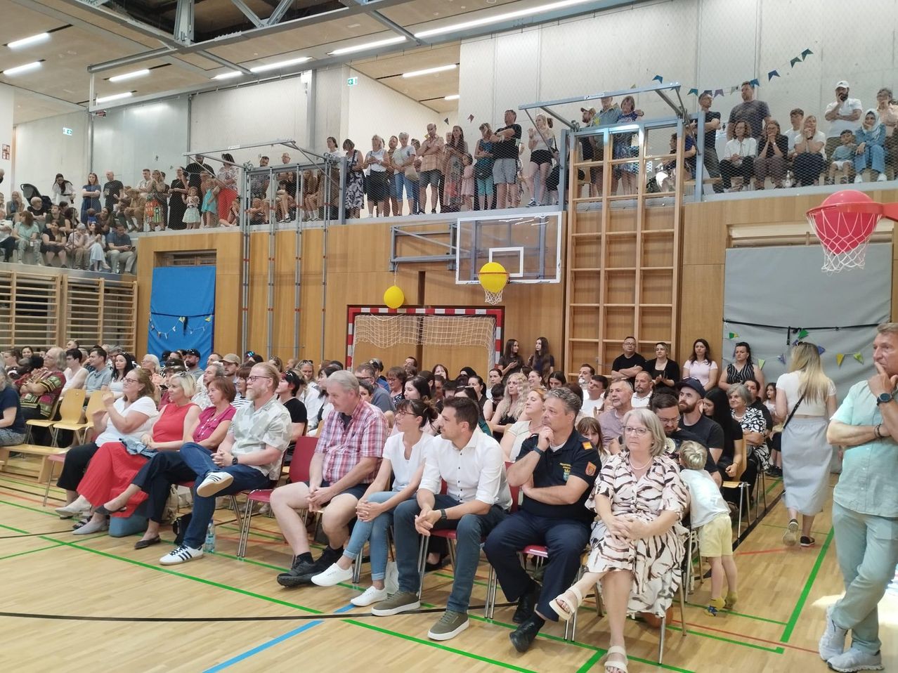 Eine große Menschenmenge versammelt sich in einer Turnhalle und schaut sich ein Sportereignis an. Zuschauer sitzen auf Stühlen und Tribünen, während andere auf der Galerie stehen. Ein Basketballkorb und ein gelber Ball sind sichtbar.