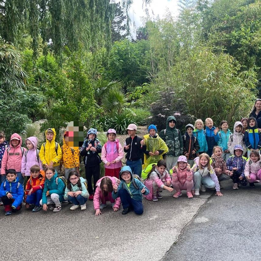 Eine Gruppe von Kindern in Regenmänteln und Mützen, einige in Hockstellung, posiert für ein Foto in einem Außenbereich mit Straße, umgeben von viel Grün.