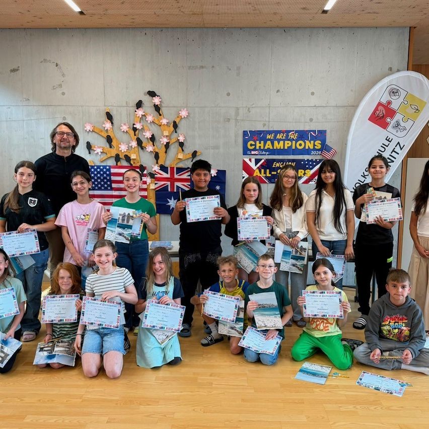 Eine Gruppe von Schülern und Lehrern posiert mit Zertifikaten, mit einer dekorierten Wand im Hintergrund.