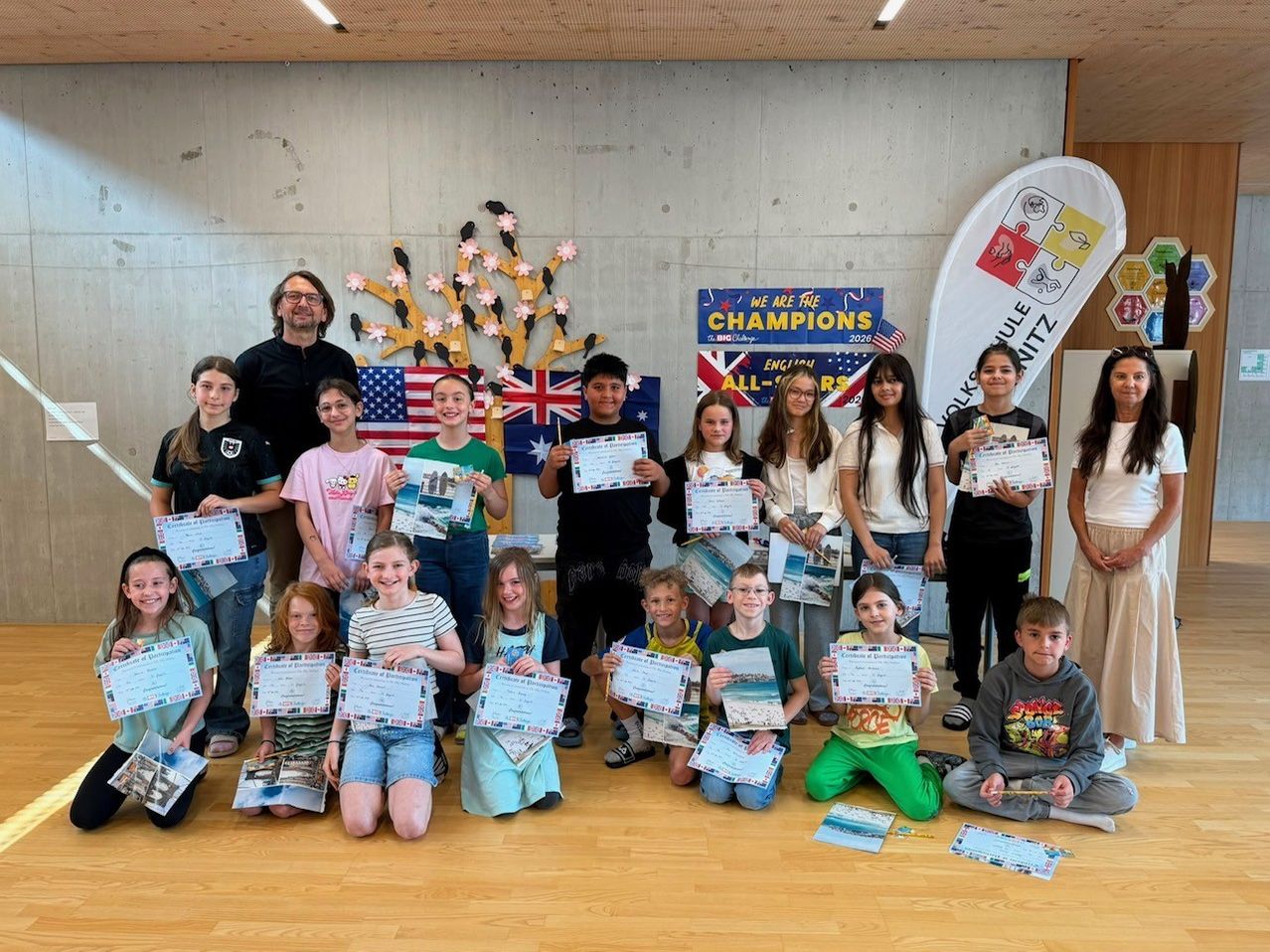 Eine Gruppe von Schülern und Lehrern posiert mit Zertifikaten, mit einer dekorierten Wand im Hintergrund.