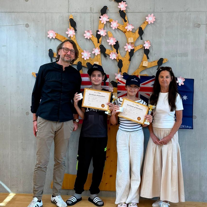 Eine vierköpfige Familie steht vor einem dekorativen Baum mit Blumen und australischen Flaggen. Sie halten alle Urkunden in der Hand. Der Mann und die beiden Jungen tragen schwarze Hemden und Hosen. Die Frau trägt ein weißes Hemd und einen Rock.