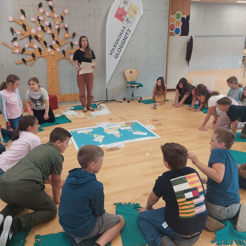 Kinder sitzen im Kreis auf dem Boden mit einer Karte. Eine Lehrerin steht mit Papier vor ihnen. Ein Baum und ein Banner sind an der Wand.