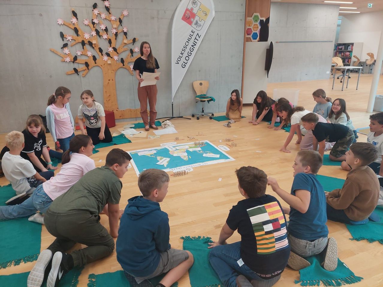 Kinder sitzen im Kreis auf dem Boden mit einer Karte. Eine Lehrerin steht mit Papier vor ihnen. Ein Baum und ein Banner sind an der Wand.