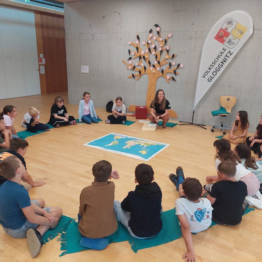 Eine Gruppe von Kindern sitzt im Kreis mit einer Frau und betrachtet eine Weltkarte auf dem Boden. Sie befinden sich in einem Klassenzimmer mit einer hölzernen Baumdekoration und einem Banner.