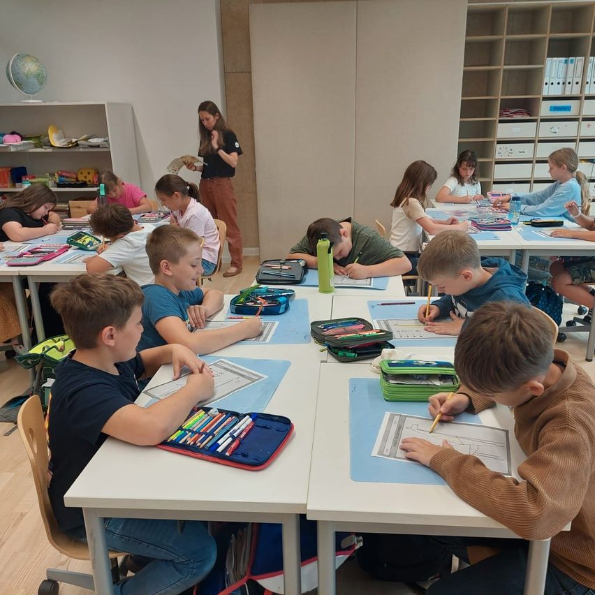 Ein Klassenzimmer voller Kinder, die an Schreibtischen sitzen. Die Lehrerin steht vorne, und die Schüler arbeiten an ihren Aufgaben. Verschiedene Schulsachen sind auf den Schreibtischen verstreut.