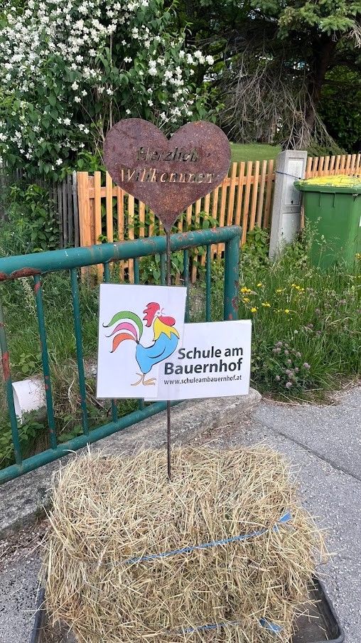 Ein herzförmiges Schild mit deutschem Text und einem Hahnenlogo steht auf einem Heuballen. Dahinter befindet sich ein grüner Metallzaun mit einem hölzernen Zaun darüber.