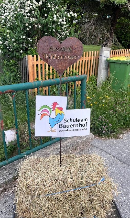 Ein herzförmiges Schild mit deutschem Text und einem Hahnenlogo steht auf einem Heuballen. Dahinter befindet sich ein grüner Metallzaun mit einem hölzernen Zaun darüber.