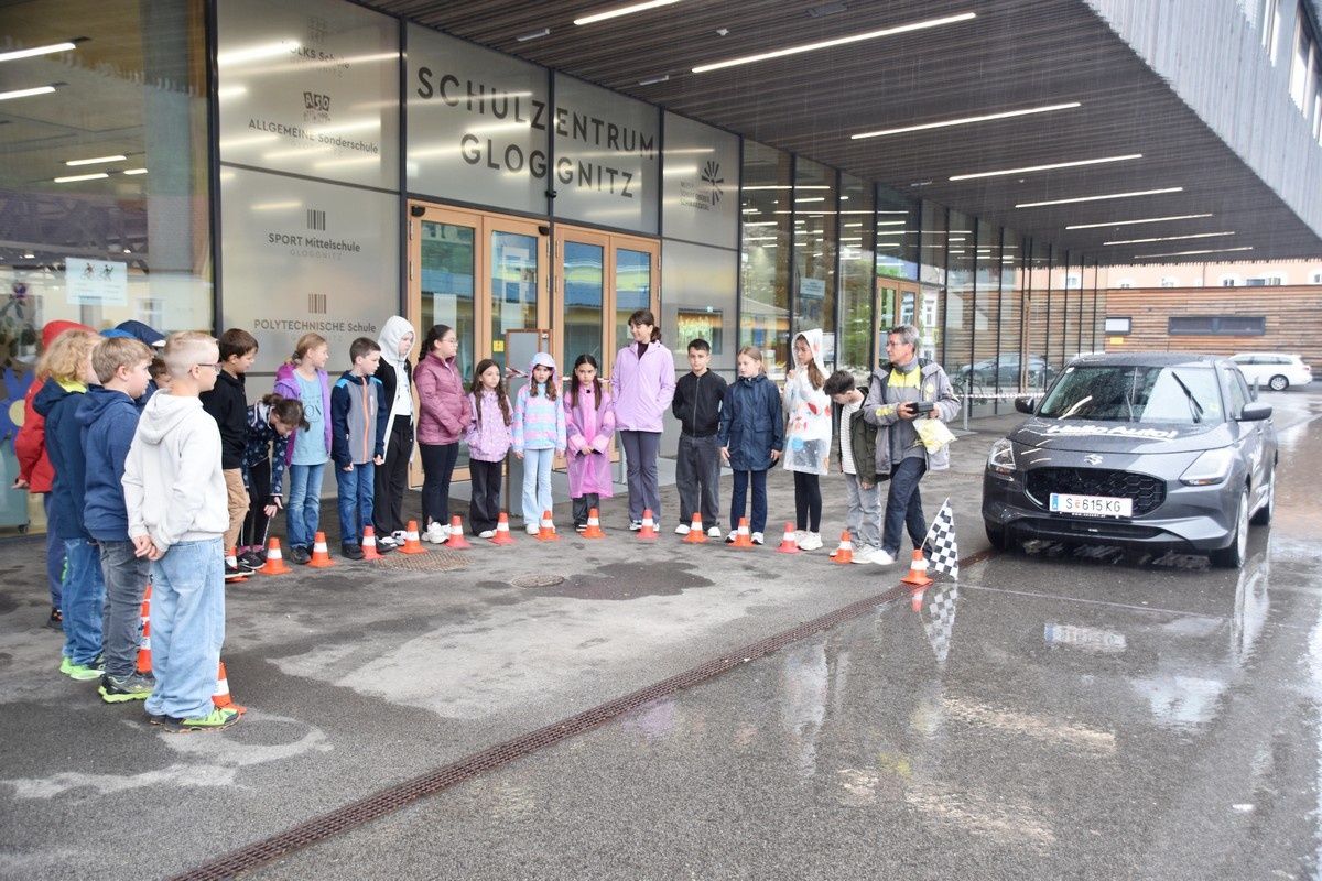 Eine Gruppe von Kindern in Jacken steht in einer Reihe, wahrscheinlich für ein Rennen, mit Verkehrskegeln und einer gemusterten Flagge. Hinter ihnen befindet sich ein Gebäude mit Schildern wie 'SCHULZENTRIUM' und 'SPORT MITTELSCHULE'.