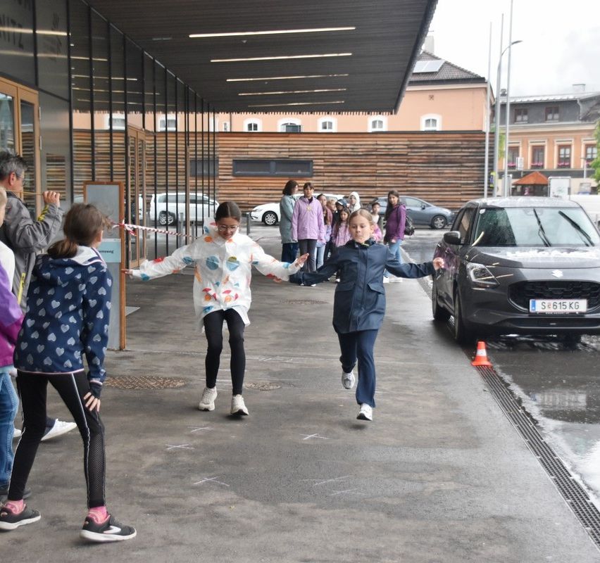 Kinder laufen über einen nassen Bürgersteig, mit einem geparkten Auto und einem orangefarbenen Kegel in der Nähe. Dahinter sind eine Reihe von Menschen und geparkte Autos zu sehen.