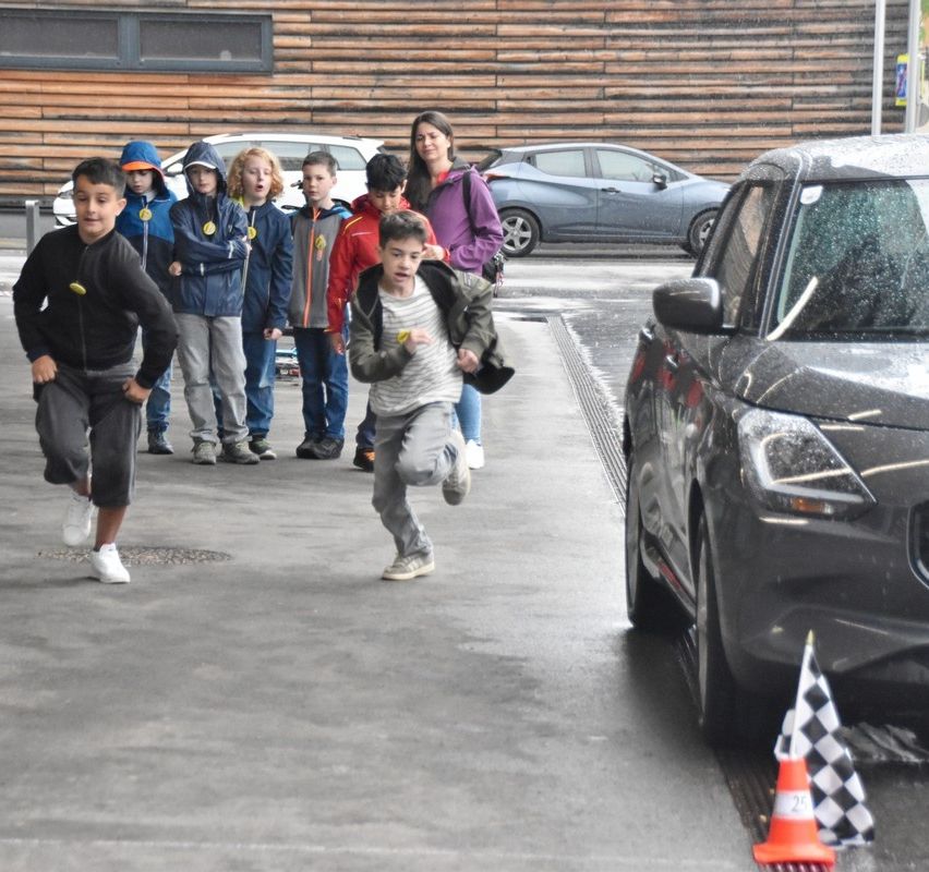 Mehrere Kinder stehen in der Nähe eines Gebäudes und einer Straße mit Autos. Ein Kind läuft, während andere zusehen. Ein Verkehrskegel befindet sich in der Nähe.