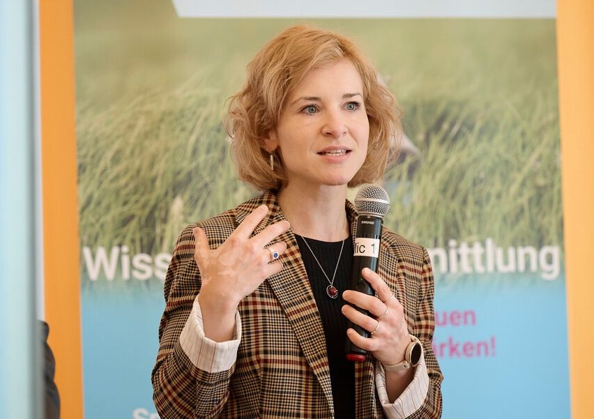 Eine Frau spricht in ein Mikrofon. Sie trägt eine karierte Jacke, ein schwarzes Oberteil und hat kurze blonde Haare. Hinter ihr ist ein Schild mit dem Wort 'Wissenschaft'.