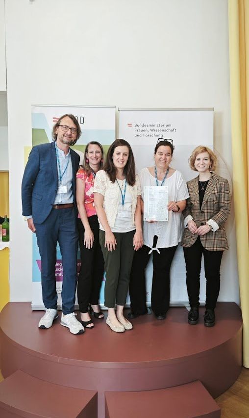Fünf Personen, darunter ein Mann und vier Frauen, stehen auf einer Bühne und halten ein Zertifikat. Sie lächeln und posieren für ein Foto. Dahinter befinden sich zwei Banner mit Text und Logos.