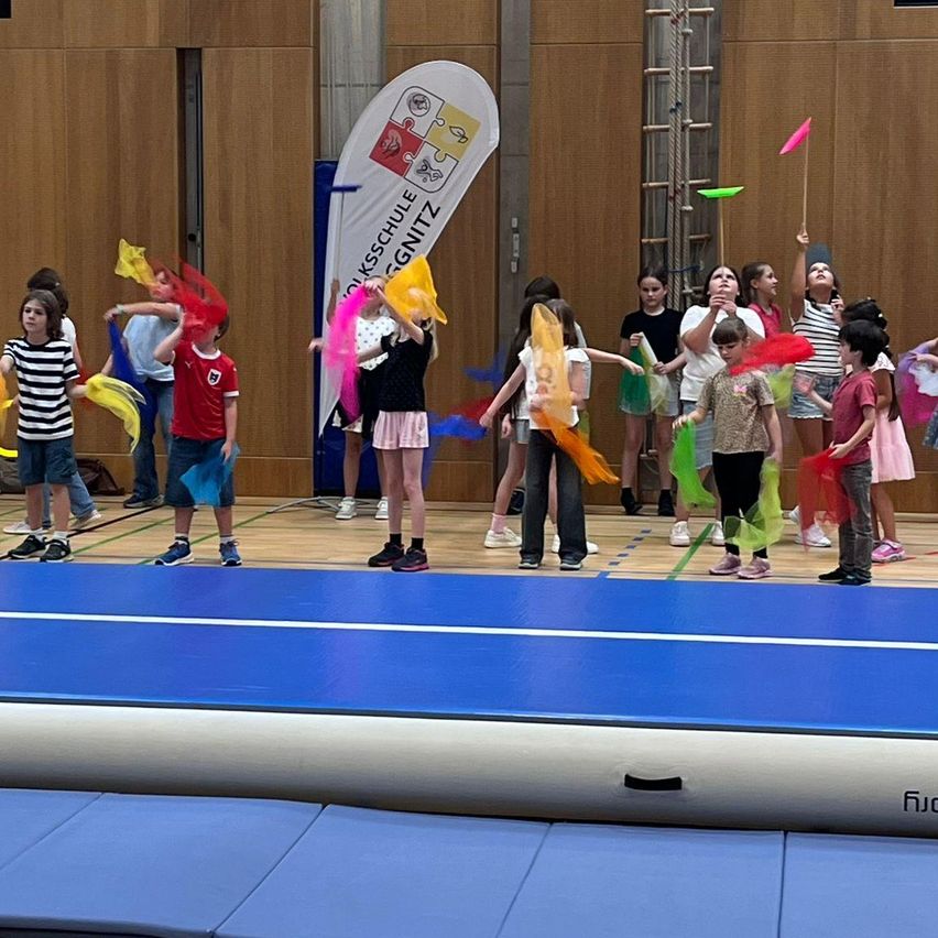 Kinder halten bunte Bänder und spielen damit in einer Turnhalle. Ein Banner mit Text ist an der Wand.