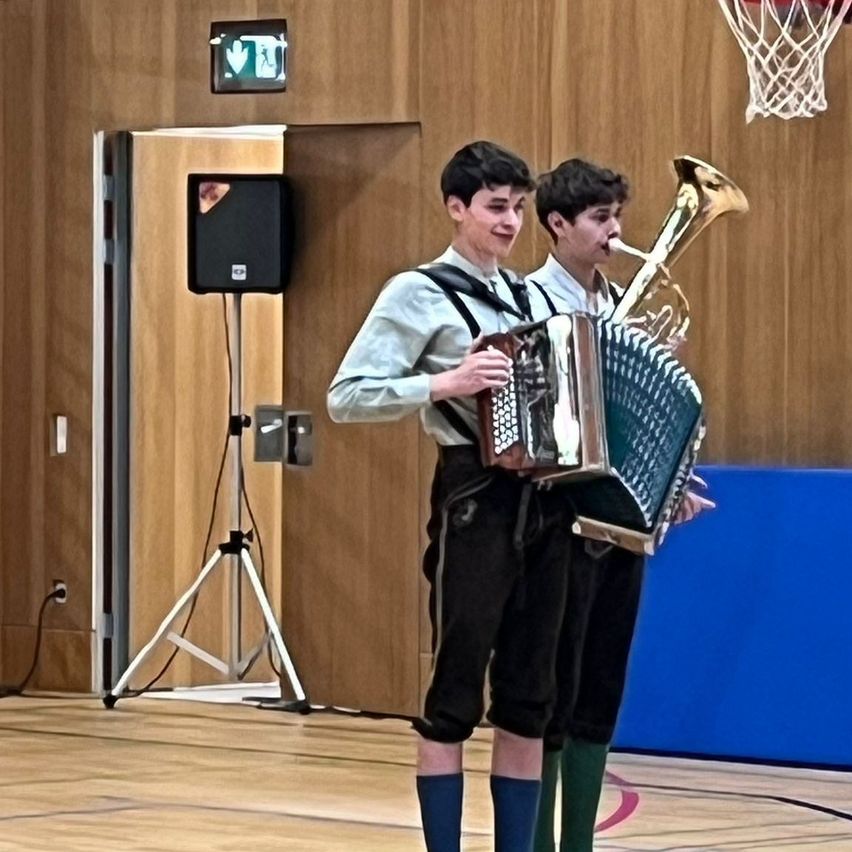 Zwei Männer spielen Musikinstrumente in einem Raum. Einer hält ein Akkordeon, der andere eine Tuba. Ein Lautsprecher steht auf einem Stativ in der Nähe, und ein Basketballkorb ist an der Wand montiert.