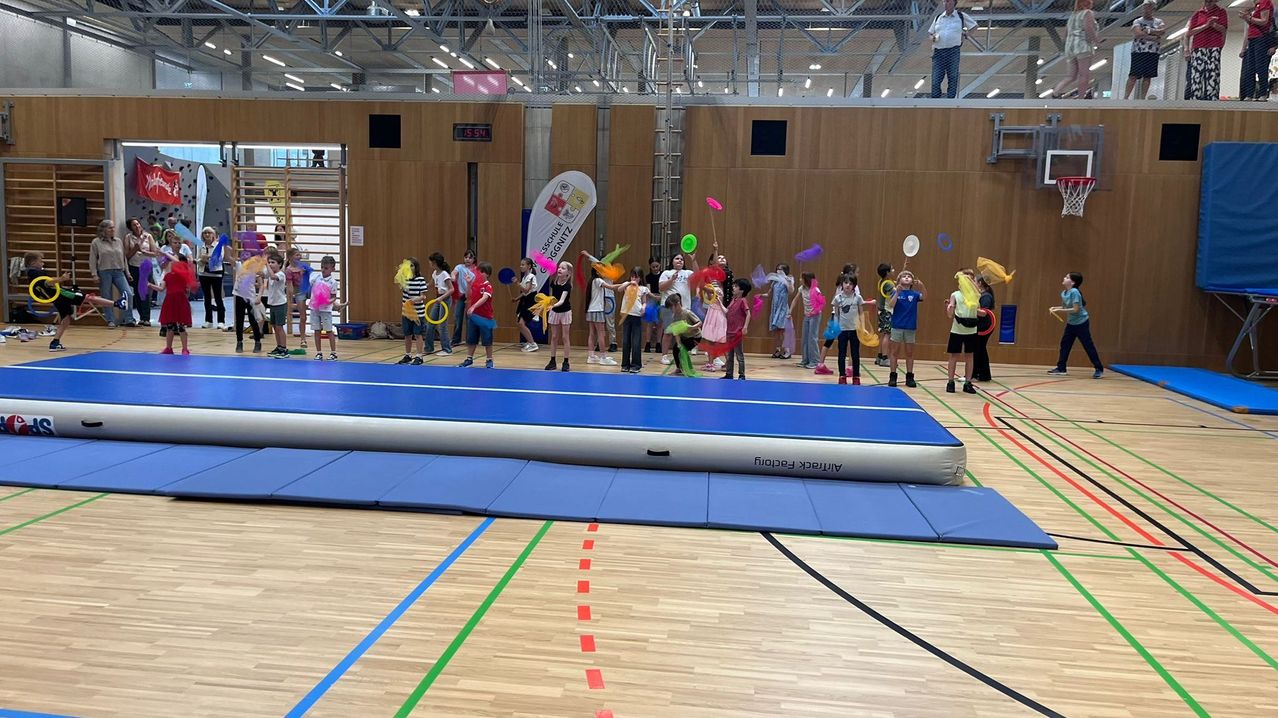 Eine Gruppe von Kindern spielt mit bunten Frisbees und Spielzeug in einer Turnhalle. Auf dem Boden befindet sich eine große aufblasbare blaue Matte.