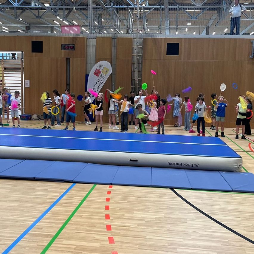 Eine Gruppe von Kindern spielt mit bunten Frisbees und Spielzeug in einer Turnhalle. Auf dem Boden befindet sich eine große aufblasbare blaue Matte.