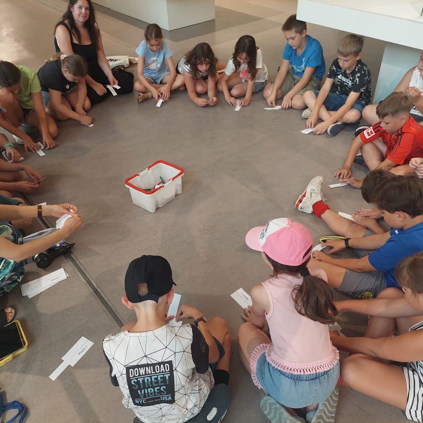 Eine Gruppe von Kindern und Erwachsenen sitzt im Kreis auf dem Boden, hält Papiere in der Hand, mit einer roten Box in der Mitte. Sie scheinen an einer Aktivität beteiligt zu sein.