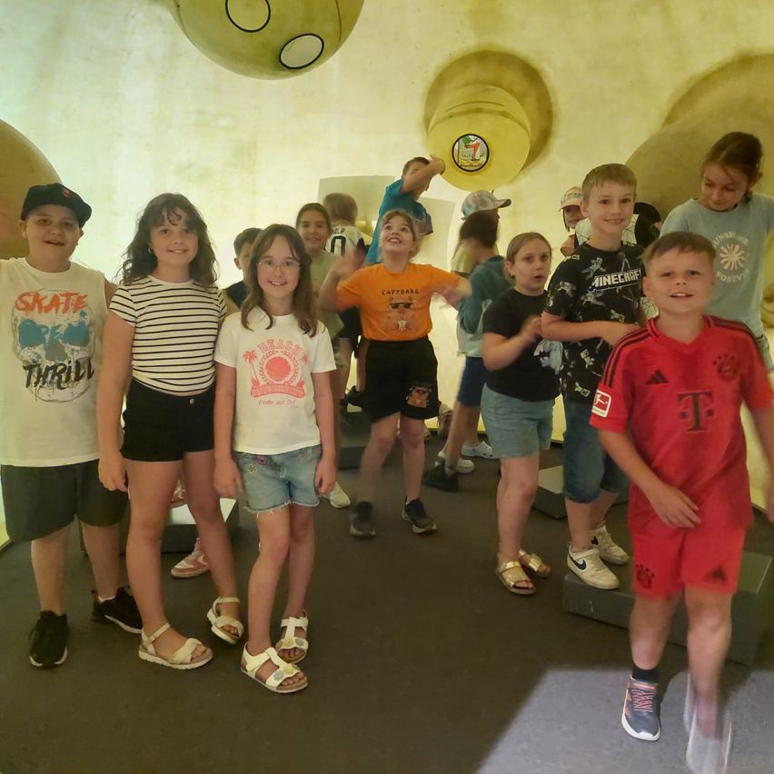 Eine Gruppe von Kindern in einem Raum mit gelben und orangefarbenen Wänden. Sie stehen und posieren für ein Foto, tragen Freizeitkleidung. Der Raum hat ein einzigartiges Design mit runden Strukturen.
