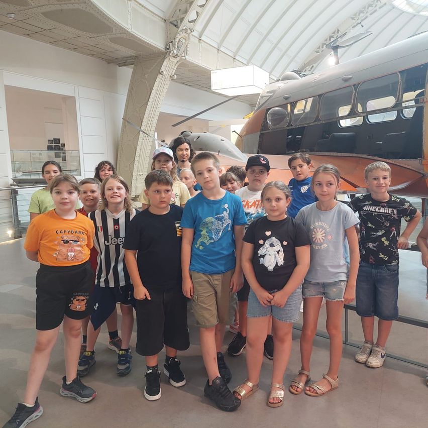 Eine Gruppe von Kindern steht zusammen vor einem Flugzeug in einer Ausstellung. Sie tragen lässige Sommerkleidung.