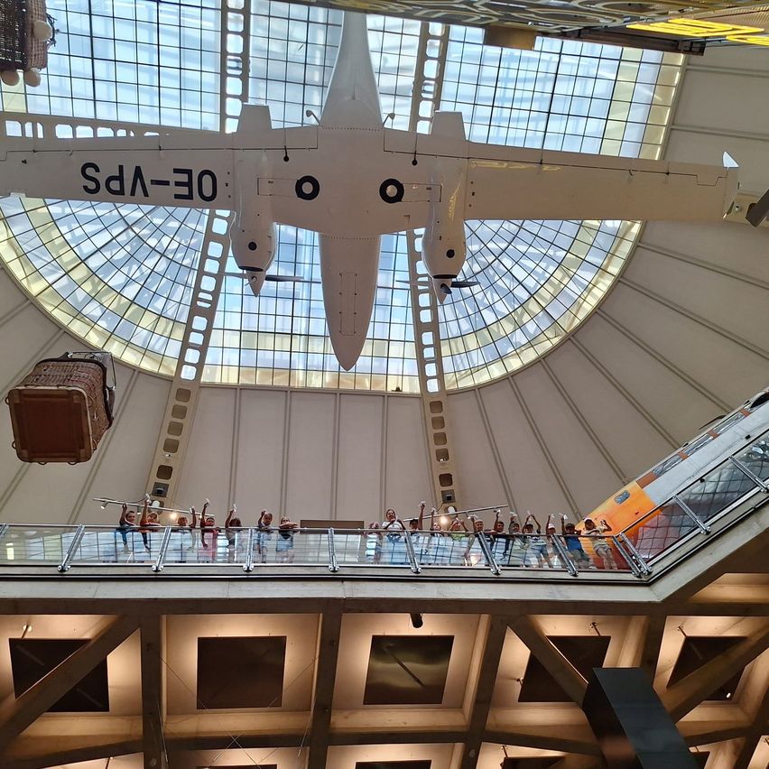 Eine Gruppe von Menschen steht auf einem Balkon unter einer großen Glaskuppel, mit einem Flugzeug, das von der Decke hängt. Das Flugzeug hat den Text '03-DA' darauf.