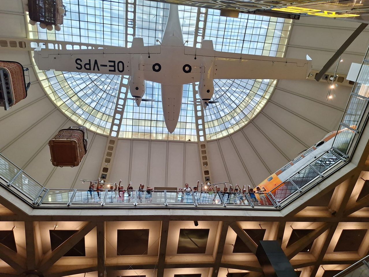 Eine Gruppe von Menschen steht auf einem Balkon unter einer großen Glaskuppel, mit einem Flugzeug, das von der Decke hängt. Das Flugzeug hat den Text '03-DA' darauf.