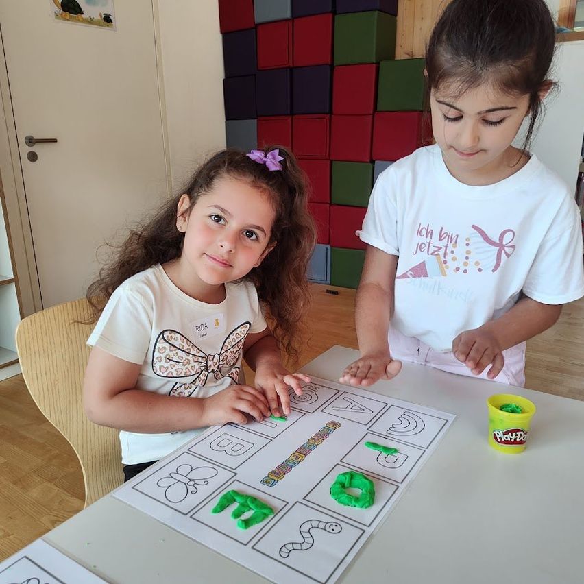 Zwei junge Mädchen in einem Klassenzimmer verwenden Play-Doh, um Buchstaben zu erstellen. Ein Mädchen trägt einen Bogen im Haar.