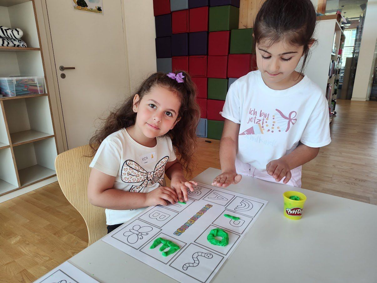 Zwei junge Mädchen in einem Klassenzimmer verwenden Play-Doh, um Buchstaben zu erstellen. Ein Mädchen trägt einen Bogen im Haar.
