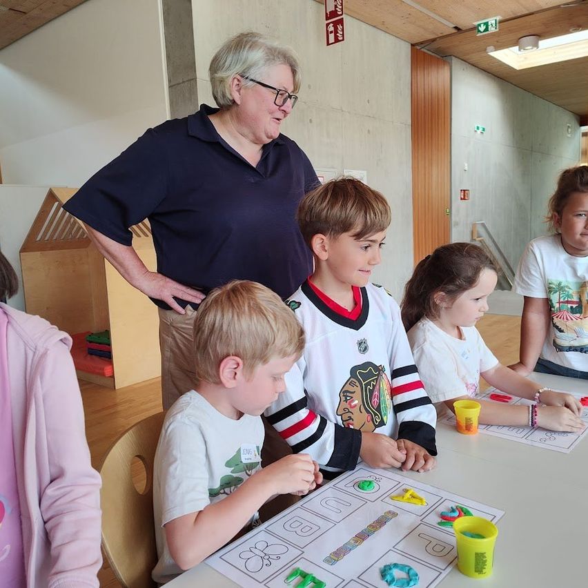 Vier Kinder und ein Erwachsener befinden sich in einem Klassenzimmer. Drei der Kinder spielen mit einem Brettspiel. Die Frau trägt eine Brille und ein blaues Hemd.