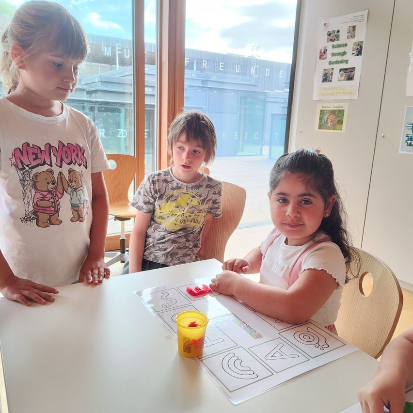 Drei Kinder sitzen an einem Tisch und beschäftigen sich mit einer Aktivität. Das Mädchen in der Mitte spielt mit kleinen Objekten auf einem Arbeitsblatt. Ein weiteres Mädchen schaut zu, während ein drittes von der Seite beobachtet. Der Raum hat eine Glaswand, die einen Blick auf ein Gebäude bietet.