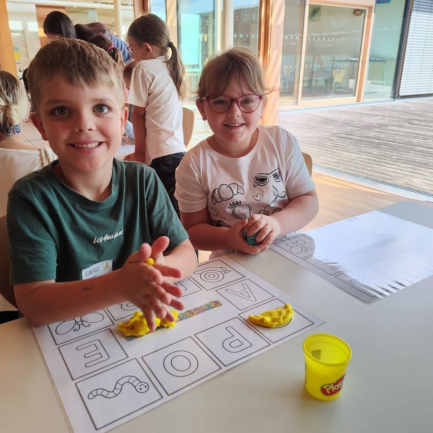 Zwei Kinder sitzen mit Play-Doh und einem Arbeitsblatt am Tisch und lächeln. Das Kind in Grün formt Buchstaben, während das Kind in Weiß zusieht. Im Hintergrund sitzen andere Kinder, und es gibt einen Blick auf einen Außenbereich.