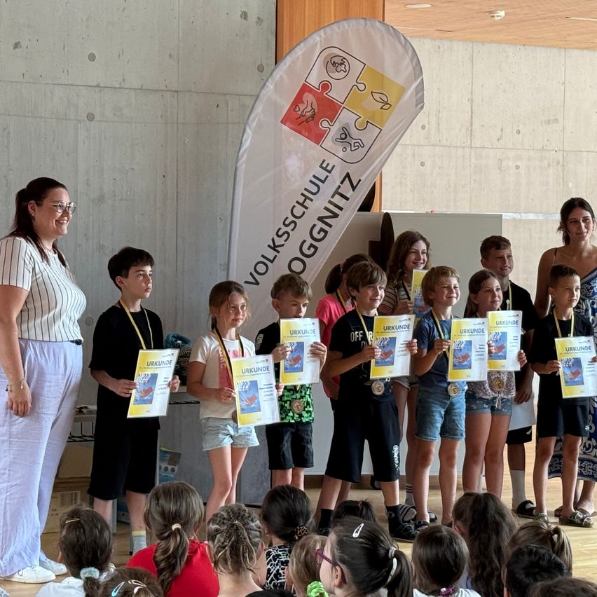 Kinder, die Urkunden halten, stehen auf der Bühne mit Erwachsenen bei einer Veranstaltung für die Volksschule Oggnitz. Eine Frau mit Brille steht vorne.