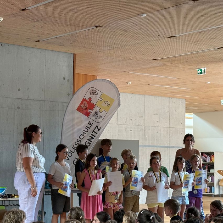 Eine Gruppe von Kindern und Jugendlichen steht vor einem Banner und hält Urkunden und Papiere in den Händen. Der Ort scheint ein Schulauditorium oder ein ähnlich großer Raum zu sein.