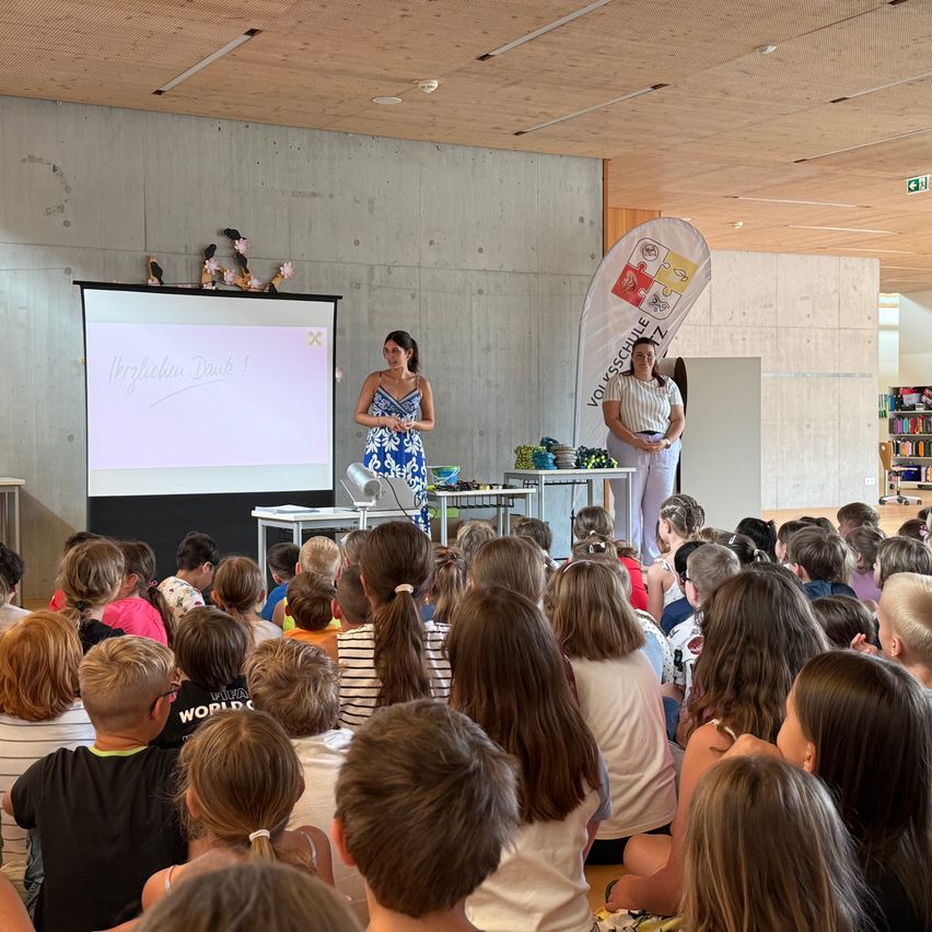 Zwei Frauen stehen an einem Podium und blicken in einen Klassenraum voller Kinder. Eine Projektionsleinwand hinter ihnen zeigt Text. Ein Banner hängt an der Wand hinter den Frauen.