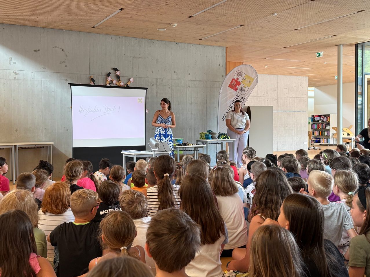 Zwei Frauen stehen an einem Podium und blicken in einen Klassenraum voller Kinder. Eine Projektionsleinwand hinter ihnen zeigt Text. Ein Banner hängt an der Wand hinter den Frauen.