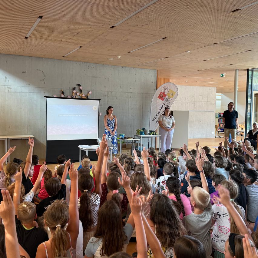 Zwei Frauen stehen an der Vorderseite eines Raumes und präsentieren vor einer Klasse von Kindern, die ihre Hände gehoben haben. Hinter den Frauen befindet sich eine Projektionsleinwand und ein Banner mit Text.