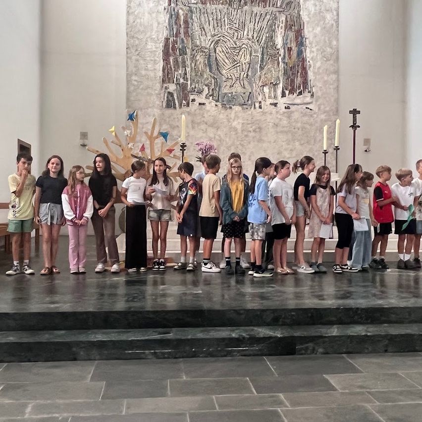 Eine Gruppe von Kindern steht auf einer Bühne vor einem großen Gemälde in einer Kirche. Einige tragen Turnschuhe, andere Sandalen. Kerzen sind in Haltern hinter ihnen.