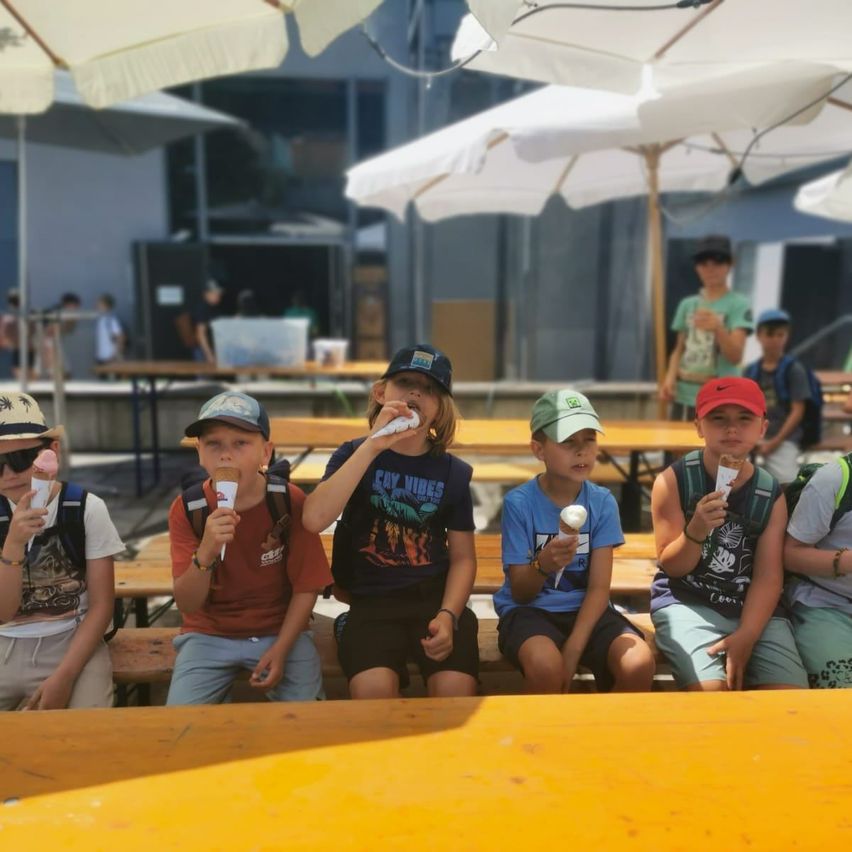 Eine Gruppe von Kindern sitzt auf Bänken und genießt Eiscreme unter weißen Schirmen. Sie tragen Mützen und Rucksäcke, mit Tischen und weiteren Kindern im Hintergrund.