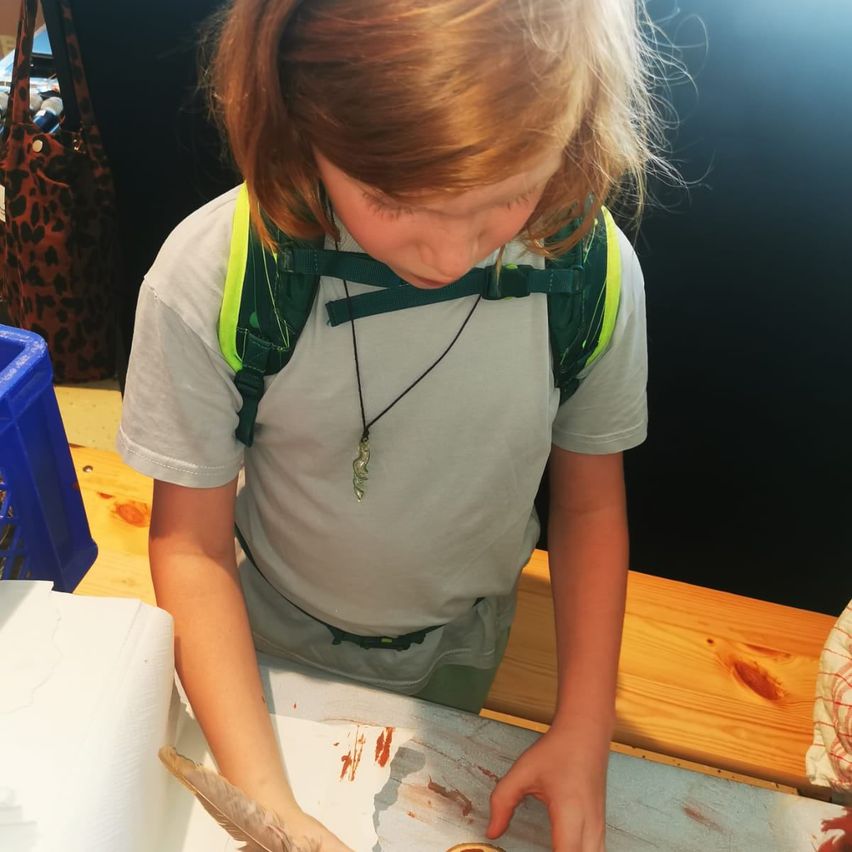 Ein kleines Mädchen in einem grauen T-Shirt und einem grünen Rucksack konzentriert sich auf ein Bastelprojekt. Sie hat eine Feder in der Hand und ihre Hände liegen auf einer weißen Oberfläche mit roten Markierungen.