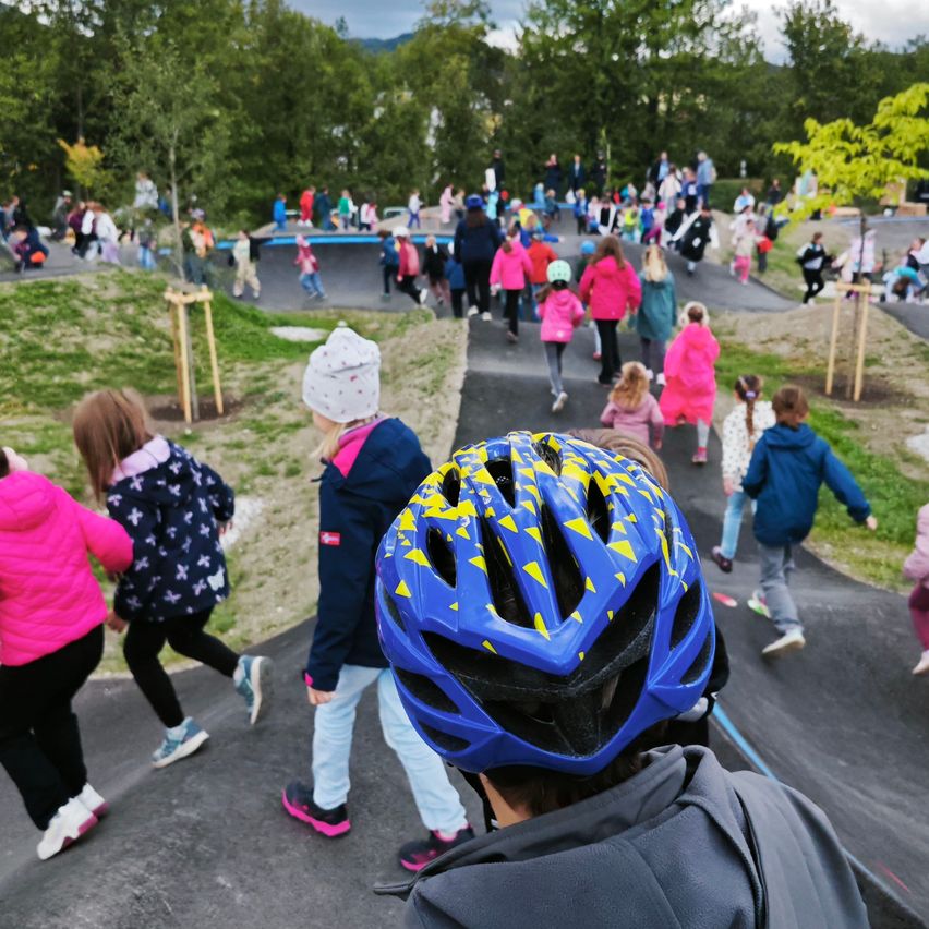 Kinder in bunten Jacken gehen auf einem gepflasterten Weg in einem Park, einige tragen Helme. Bäume und Grün umgeben sie.
