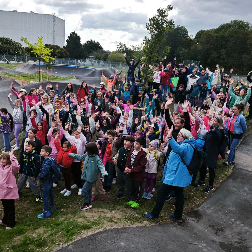 Eine große Gruppe von Kindern und Erwachsenen versammelt sich in einem Park, einige winken und lächeln, mit einem Baum und einem Skatepark im Hintergrund.