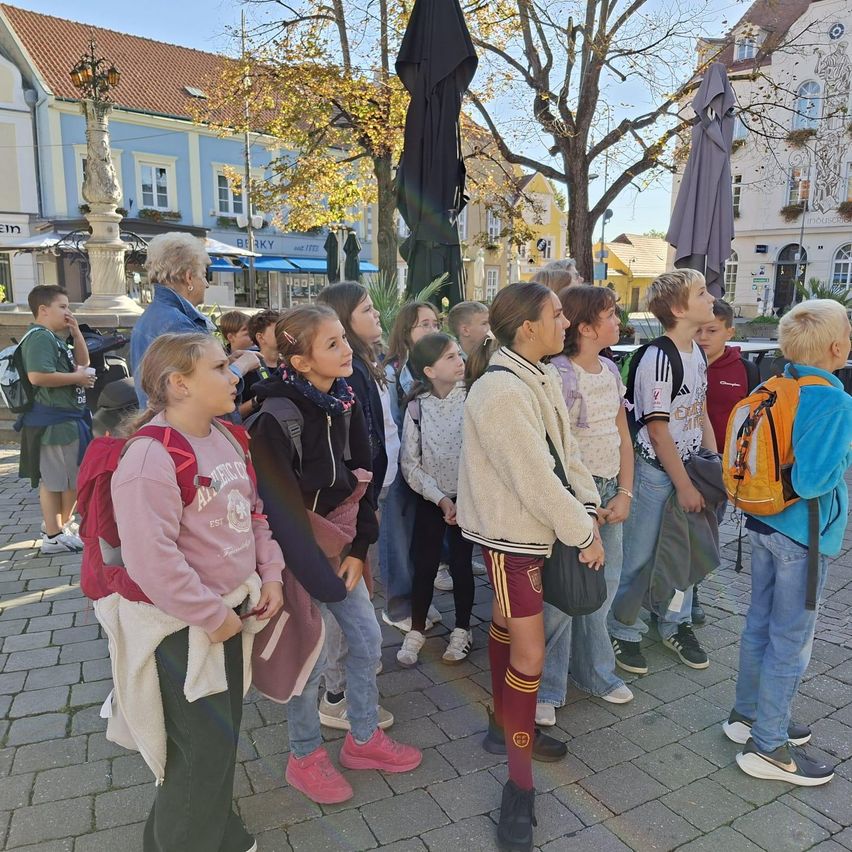Eine Gruppe von Kindern und Erwachsenen steht auf einem Stadtplatz. Sie tragen Rucksäcke und Turnschuhe.