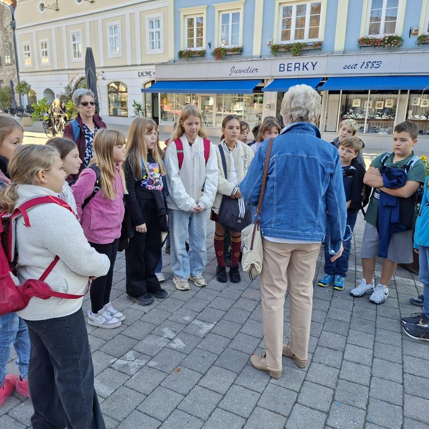 Eine Gruppe von Kindern und einem Erwachsenen stehen vor einem Gebäude. Der Erwachsene spricht mit den Kindern. Die Kinder tragen Rucksäcke und Turnschuhe.