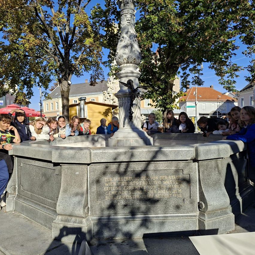 Eine Gruppe von Menschen versammelt sich um einen Brunnen auf einem Stadtplatz. Der Brunnen ist aus Stein und hat ein kunstvolles Design. Einige Leute lehnen sich an den Brunnen, während andere dahinter stehen.