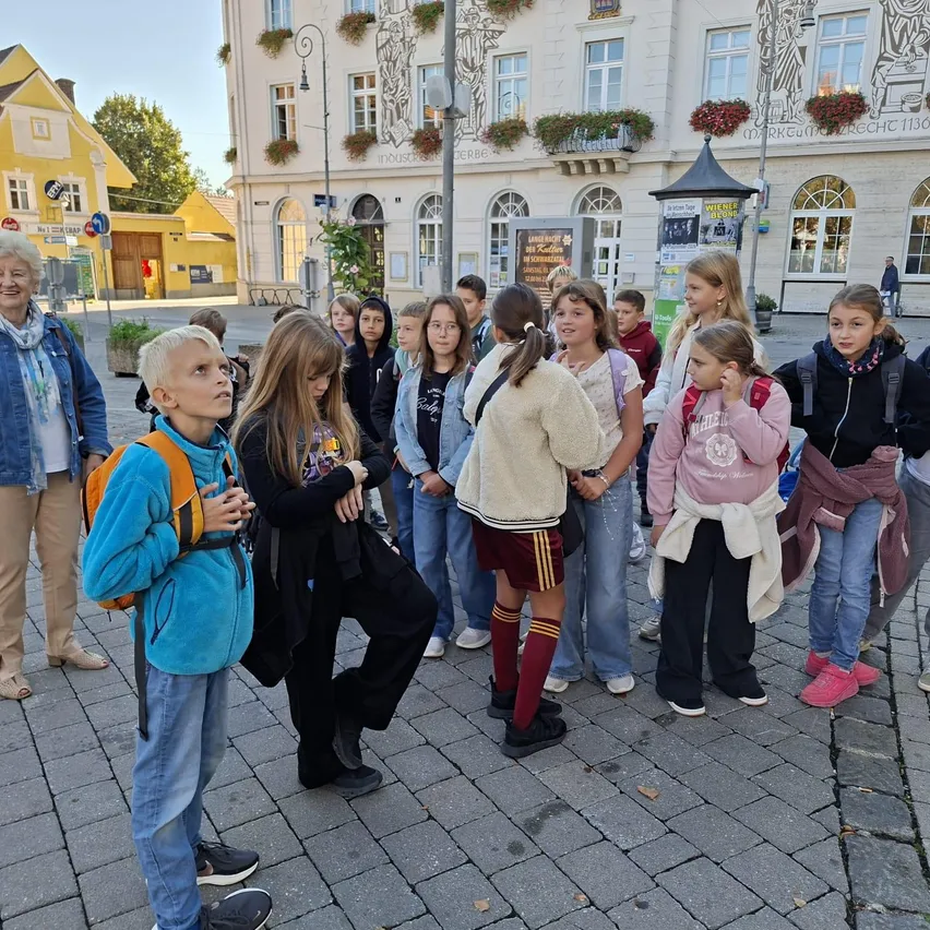 Eine Gruppe junger Schüler und eine erwachsene Frau stehen vor einem Gebäude mit gelber Fassade. Die Schüler tragen Freizeitkleidung und einige haben Rucksäcke. Die erwachsene Frau trägt eine Jeansjacke und einen Schal.