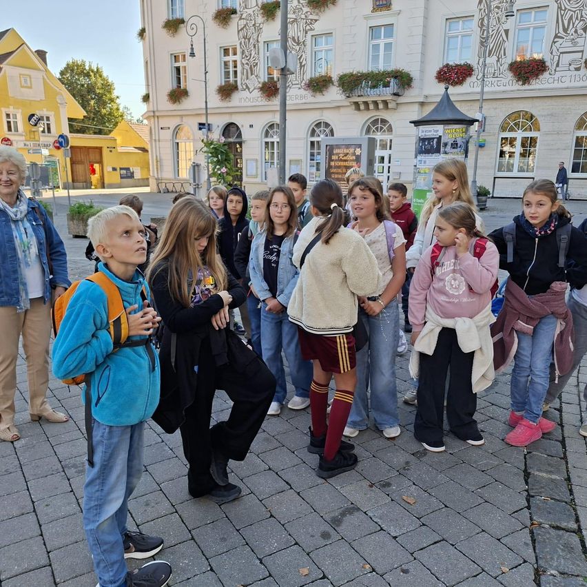 Eine Gruppe junger Schüler und eine erwachsene Frau stehen vor einem Gebäude mit gelber Fassade. Die Schüler tragen Freizeitkleidung und einige haben Rucksäcke. Die erwachsene Frau trägt eine Jeansjacke und einen Schal.