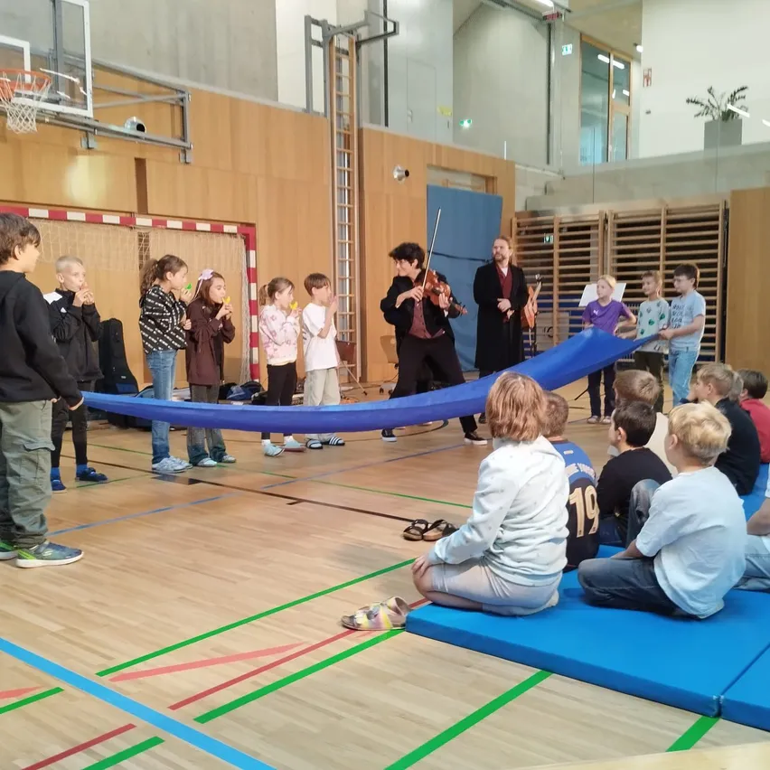 Eine Gruppe von Kindern sieht sich eine Violinenaufführung von zwei Männern in einer Turnhalle an.