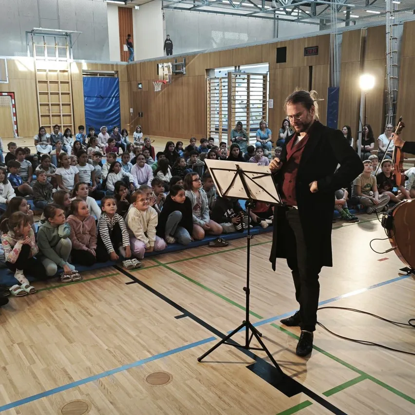 Zwei Musiker spielen auf der Bühne in einer Turnhalle. Ein Mann spielt Geige und eine Frau Gitarre. Sie stehen vor einem Notenständer und einem Publikum von Kindern, die auf dem Boden sitzen.
