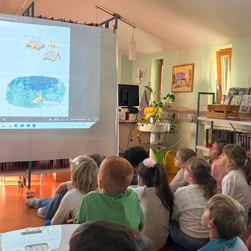 Ein Klassenzimmer mit Kindern auf dem Boden sitzend, die eine Präsentation auf einer Projektionsleinwand ansehen.